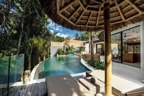 Un área de piscina con muelle de madera, sillas de descanso y una estructura cubierta de paja.
