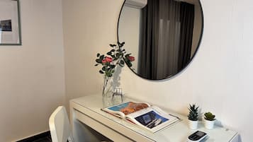 A hotel room with a white desk, a mirror, a vase with flowers, a book, a small plant, and a calculator.