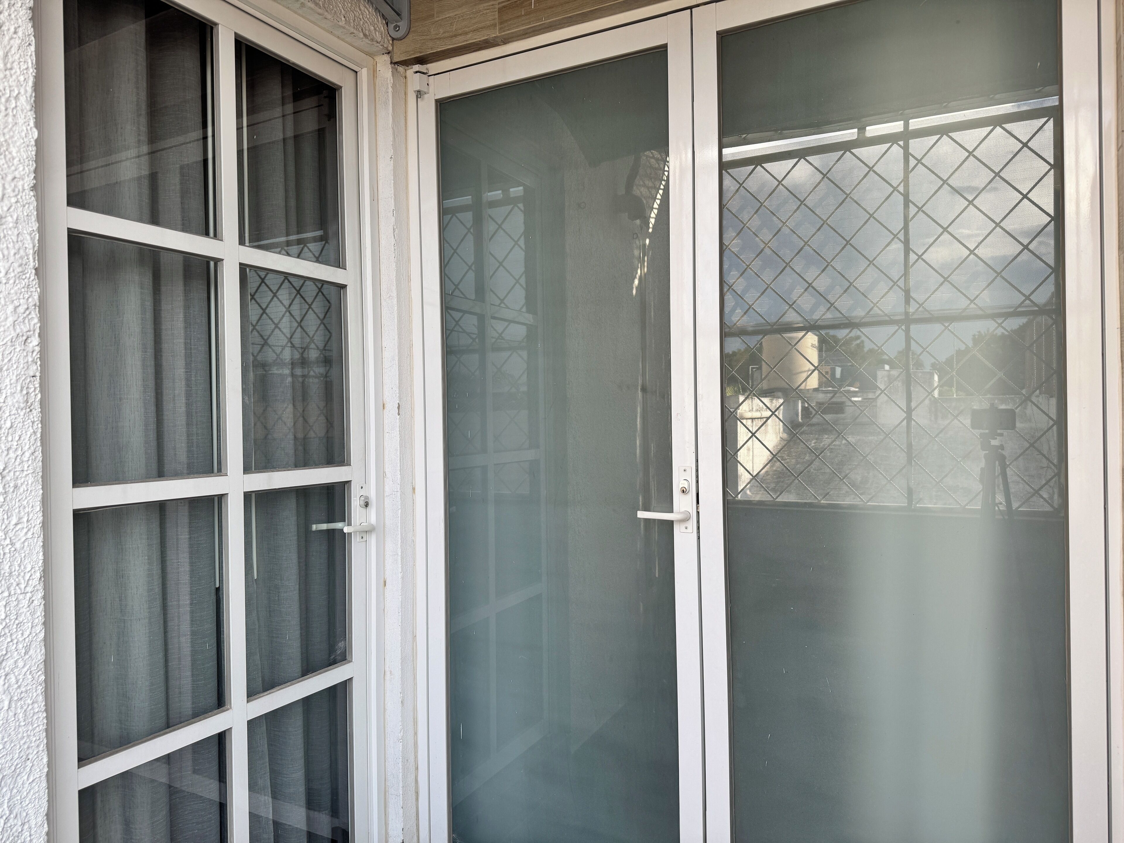 Eine Glastür mit einem Metallsicherheitsgitter, weißem Türrahmen und Blick auf einen Balkon mit Stativ und Kamera.