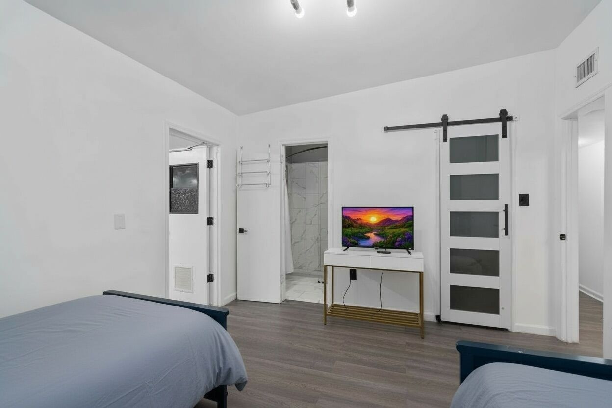 A modern bedroom with two beds, a TV on a white console, a sliding door, and a shower area.