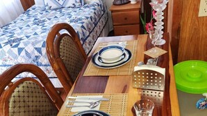 Una mesa de comedor impecablemente dispuesta con platos, cubiertos y un vaso de agua, ubicada en una habitación que incluye una cama y una mesita de noche con una lámpara y objetos decorativos.