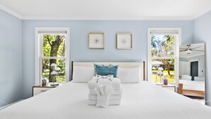 A neatly made bed with a white comforter and a folded towel, two framed wall art pieces, and a view of a tree through a window. - The Molasses Manor | Minutes from Everything (Mt Pleasant)