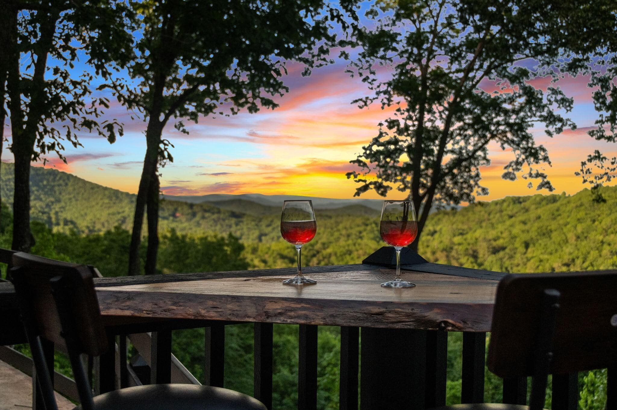 A table with two glasses of red wine, set against a backdrop of a sunset over a forested landscape.