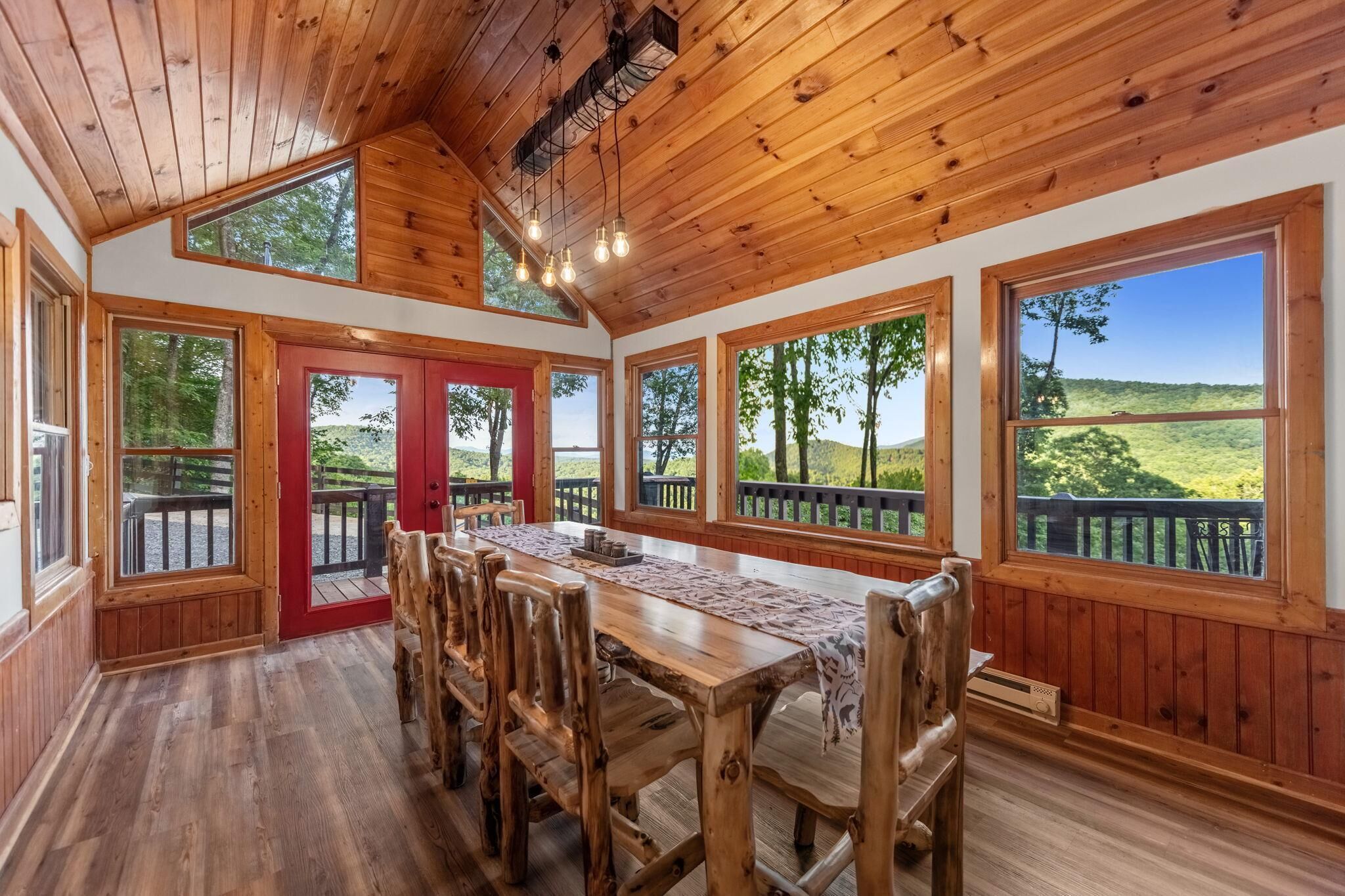 A wooden dining area with a long table and chairs, large windows with a view of a green landscape, and a red door leading outside.