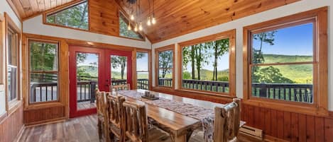A wooden dining area with a long table and chairs, large windows with a view of a green landscape, and a red door leading outside.