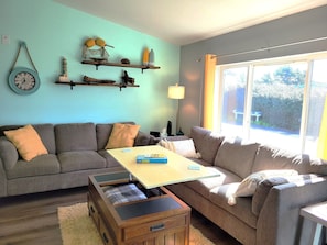 A cozy living room with a sofa, a coffee table, and a wooden chest of drawers. There are shelves with decorative items and a window with a view of greenery. - Stroll to the Beach! Warm up in the Hot Tub (Brookings)