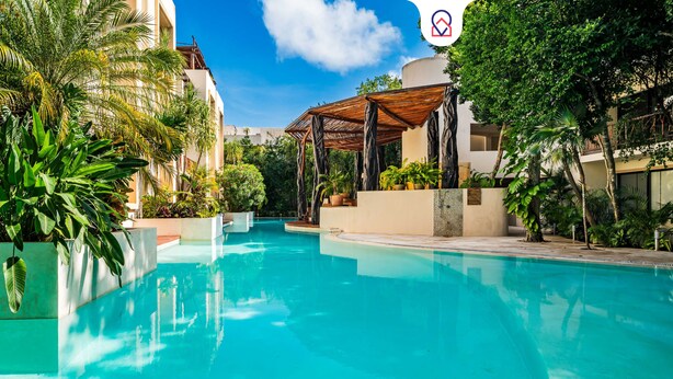 Une piscine dotée d’une structure en forme de pergola, entourée d’une végétation luxuriante et de bâtiments modernes.