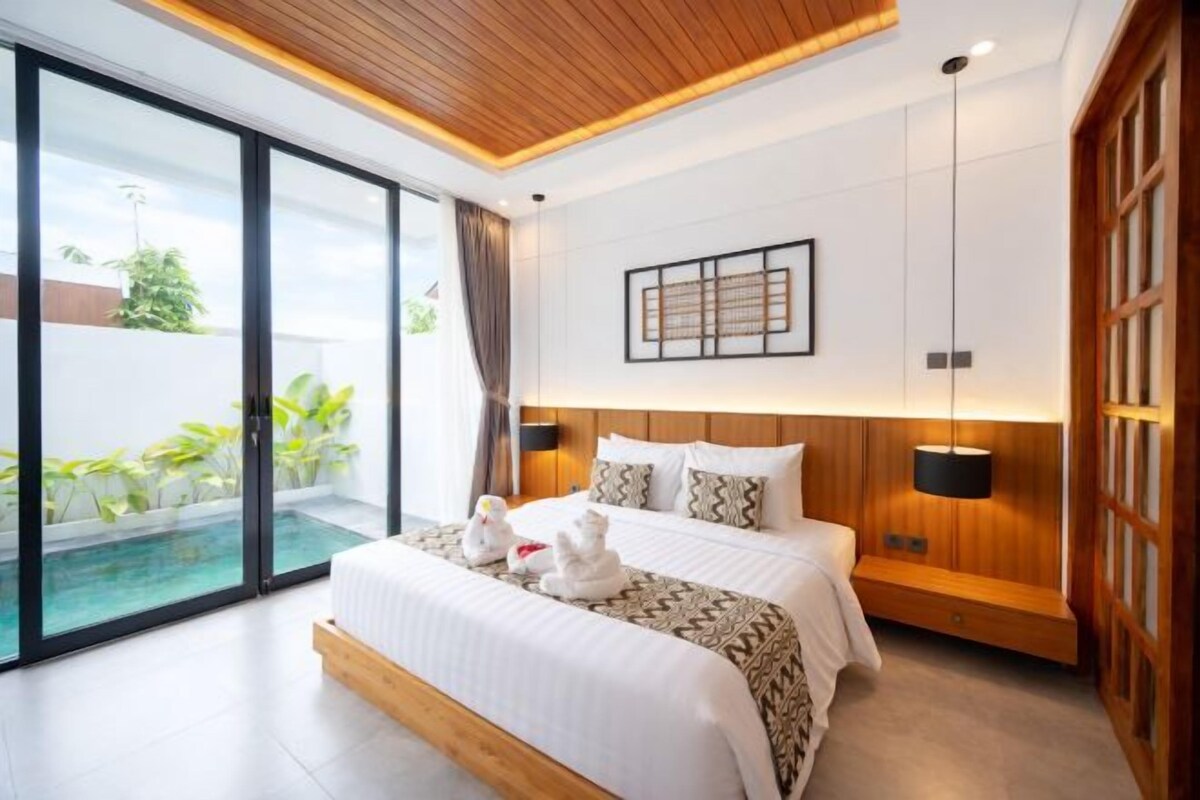 A modern hotel room with a large bed, a small seating area, a view of a swimming pool, and a sliding glass door.