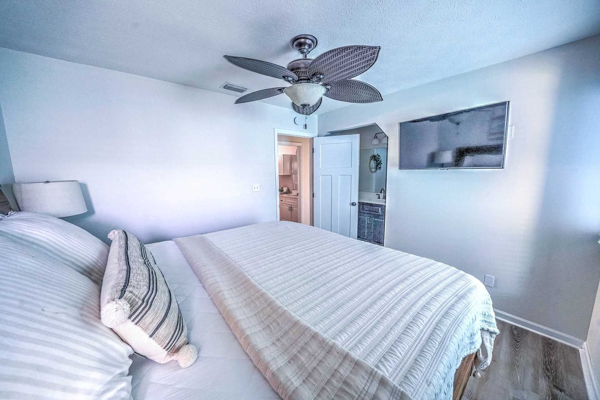 A bedroom with a bed, a ceiling fan, a nightstand, a lamp, and a view of a bathroom.