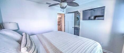 A bedroom with a bed, a ceiling fan, a nightstand, a lamp, and a view of a bathroom.