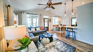 A spacious living room with a large sofa, a coffee table, and a ceiling fan. There is a kitchen area with a bar and stools. - 204 Squid Lane - Adorable Beach House! 3 Bedroom 2 baths - Golf Cart included! (Panama City Beach)