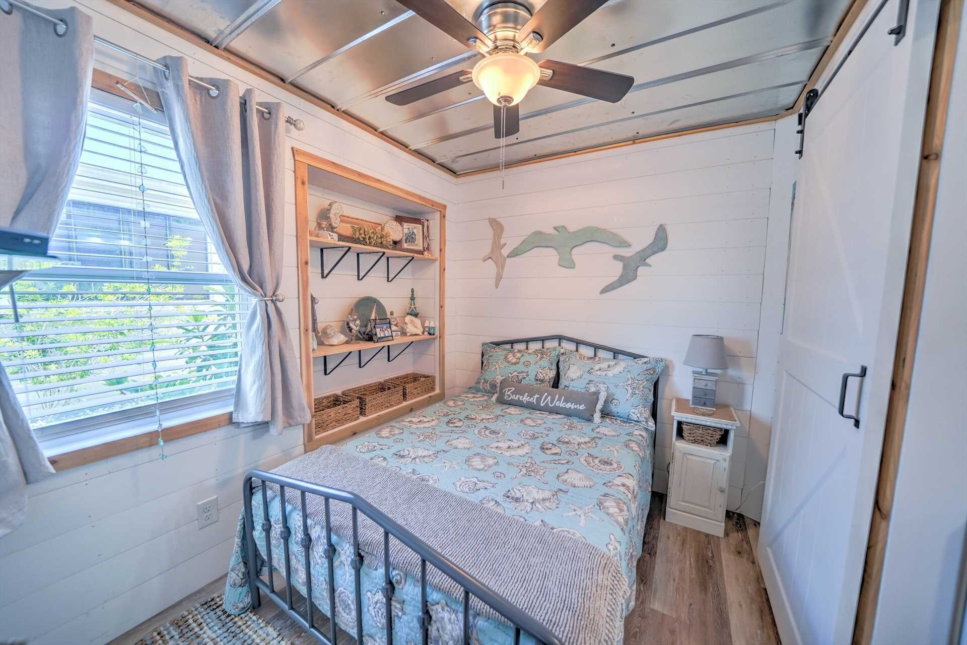 Une chambre à coucher comprenant un lit, une table de chevet, un ventilateur de plafond, une fenêtre avec des rideaux et des éléments de décoration murale.