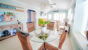 Un vaste salon doté d’une table à manger au plateau en verre, entourée de chaises en osier, d’une grande plante en pot et d’un ventilateur de plafond.