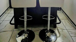 Une petite pièce moderne comprenant une table à manger et deux tabourets de bar, une plante en pot et une bibliothèque agrémentée d’objets décoratifs.