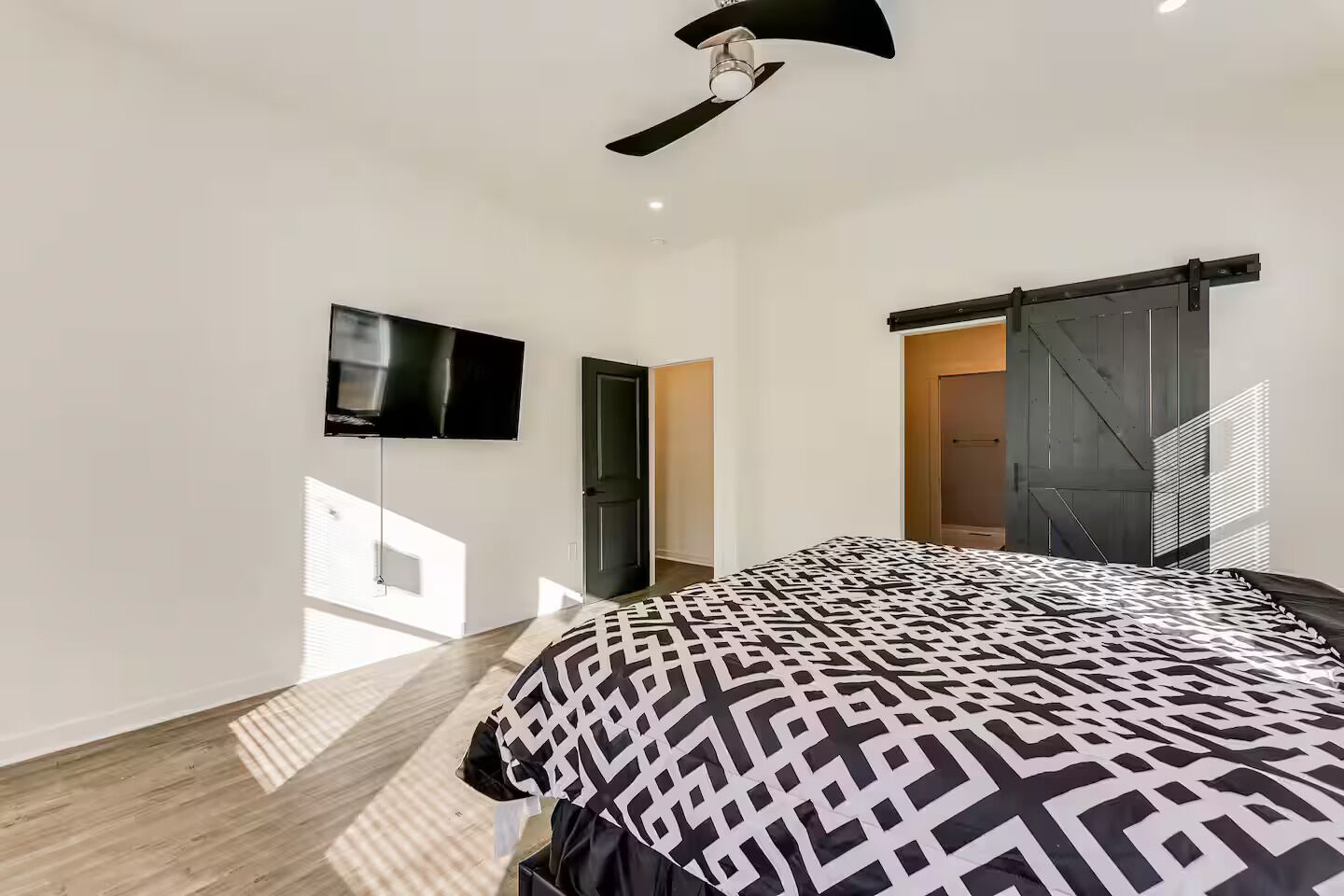 Ein modernes Schlafzimmer mit einem großen Bett, einem Deckenventilator, einem an der Wand montierten Fernseher und einer Schiebetür zu einem anderen Raum.