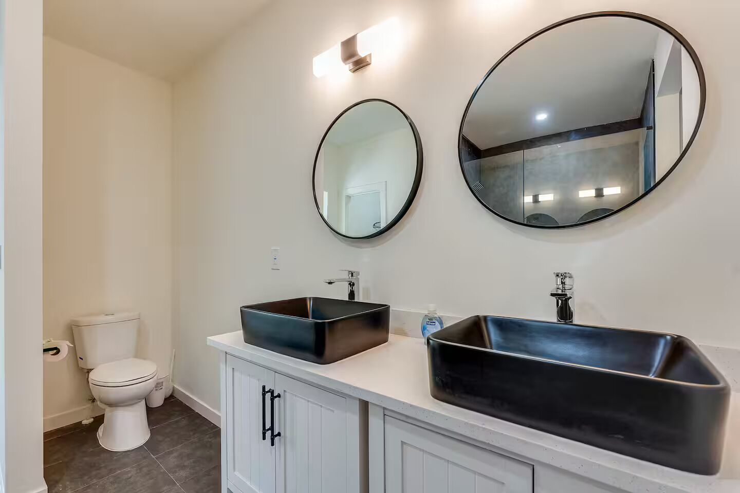 Une salle de bain avec des toilettes blanches, deux lavabos noirs et deux miroirs ronds accrochés au mur.