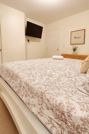 Ein Schlafzimmer mit einem zudeckenden Bett in Blumenmuster, einem an der Wand befestigten Flachbildfernseher, einer Tür und einem gerahmten Schild mit der Aufschrift 'blessed'.