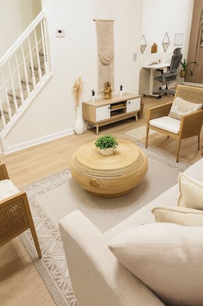 Una sala moderna con una mesa de centro redonda de madera, un sofá blanco, una silla de mimbre, un mueble de madera y una escalera.