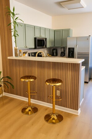 Eine moderne Küche mit grünen Schränken, einem Edelstahlkühlschrank, einer Kaffeemaschine und zwei goldenen Barhocker.