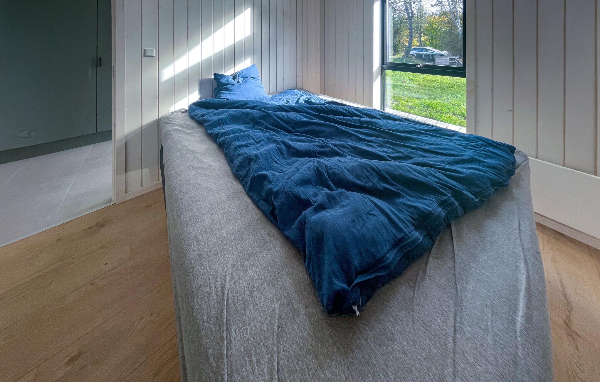 Ein Bett in einem Zimmer mit Holzböden und -wänden, ein Fenster mit Blick auf ein Auto und Grünfläche.
