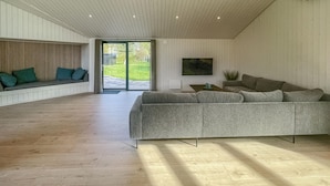 A spacious living room with a large grey sectional sofa, wooden flooring, a wooden slatted wall, and a sliding glass door leading to an outdoor area.