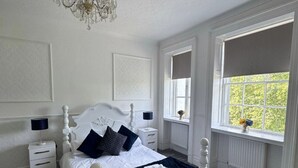 Una cama con dosel blanco, con colcha y almohadas azul oscuro, una mesita de noche blanca con una lámpara, y un ventanal con vistas a zonas verdes.