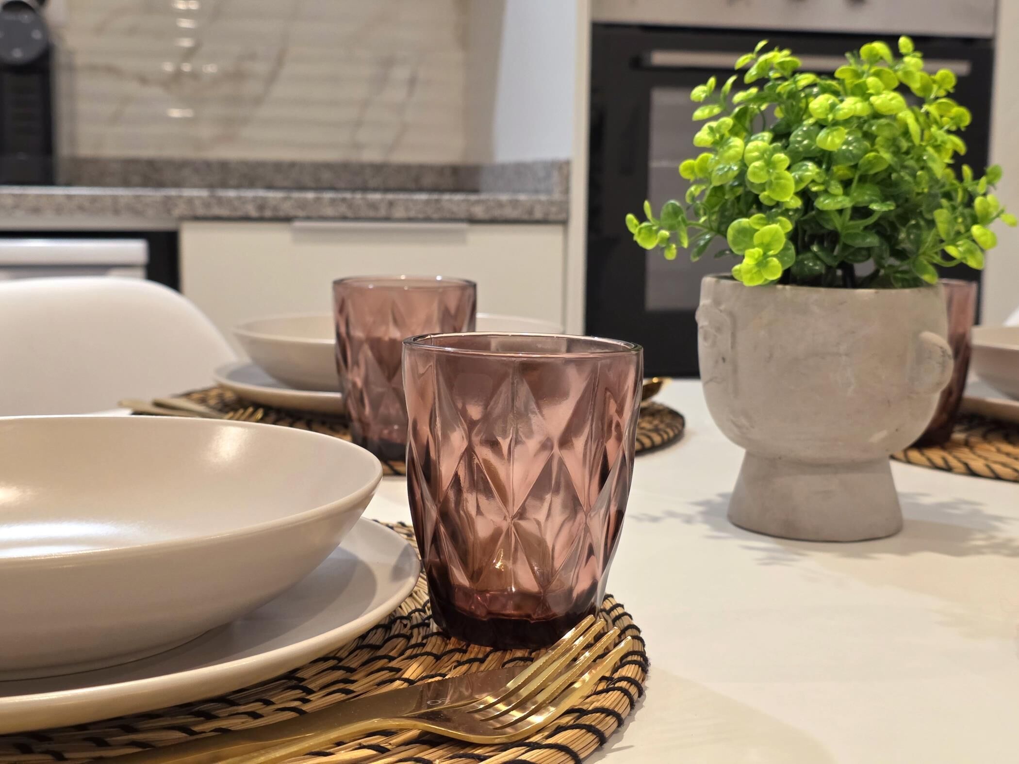 A dining table with a white plate, two pink glasses, a gold fork, and a potted plant.