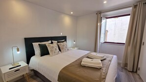 A neatly made bed with a dark headboard, white bedding, and a brown throw blanket. There are two bedside tables with lamps and a remote control on one. A window with curtains is visible, and towels are folded on the bed. - Aconchego da Vila | Olival Flat | Castelo de Vide (Castelo de Vide)