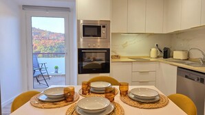 A modern kitchen with a built-in oven and microwave, a dining table set with plates and glasses, and a view of autumn foliage through the door. - Aconchego da Vila | Serra Flat | Castelo de Vide (Castelo de Vide)