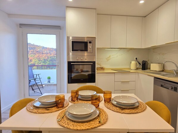 A modern kitchen with a built-in oven and microwave, a dining table set with plates and glasses, and a view of autumn foliage through the door.