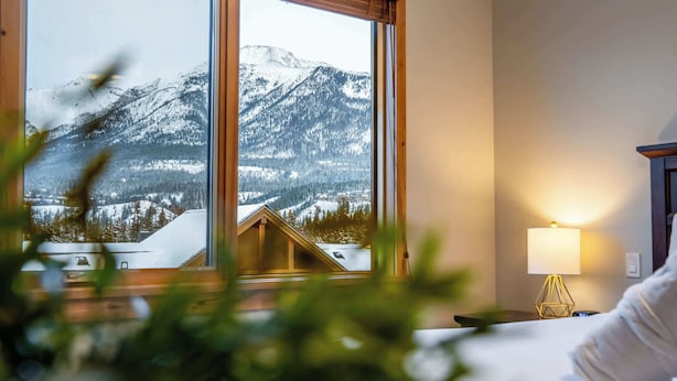 Hotellihuone, jossa on suuri ikkuna, josta avautuu näkymä lumihuippuisiin vuoriin ja mökkiin.