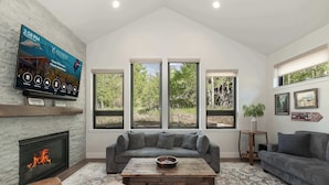 A living room with a fireplace, a large flat-screen TV, a gray sectional sofa, a wooden coffee table, and a patterned rug. - Trailside Location | Mountain Escape (Granby)