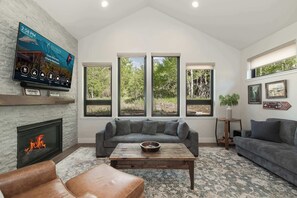 A living room with a fireplace, a large flat-screen TV, a gray sectional sofa, a wooden coffee table, and a patterned rug. - Trail Views | Hot Tub (Granby)