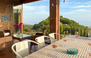 Une terrasse avec des meubles en osier, une table recouverte d’une nappe à carreaux et un verre de vin.