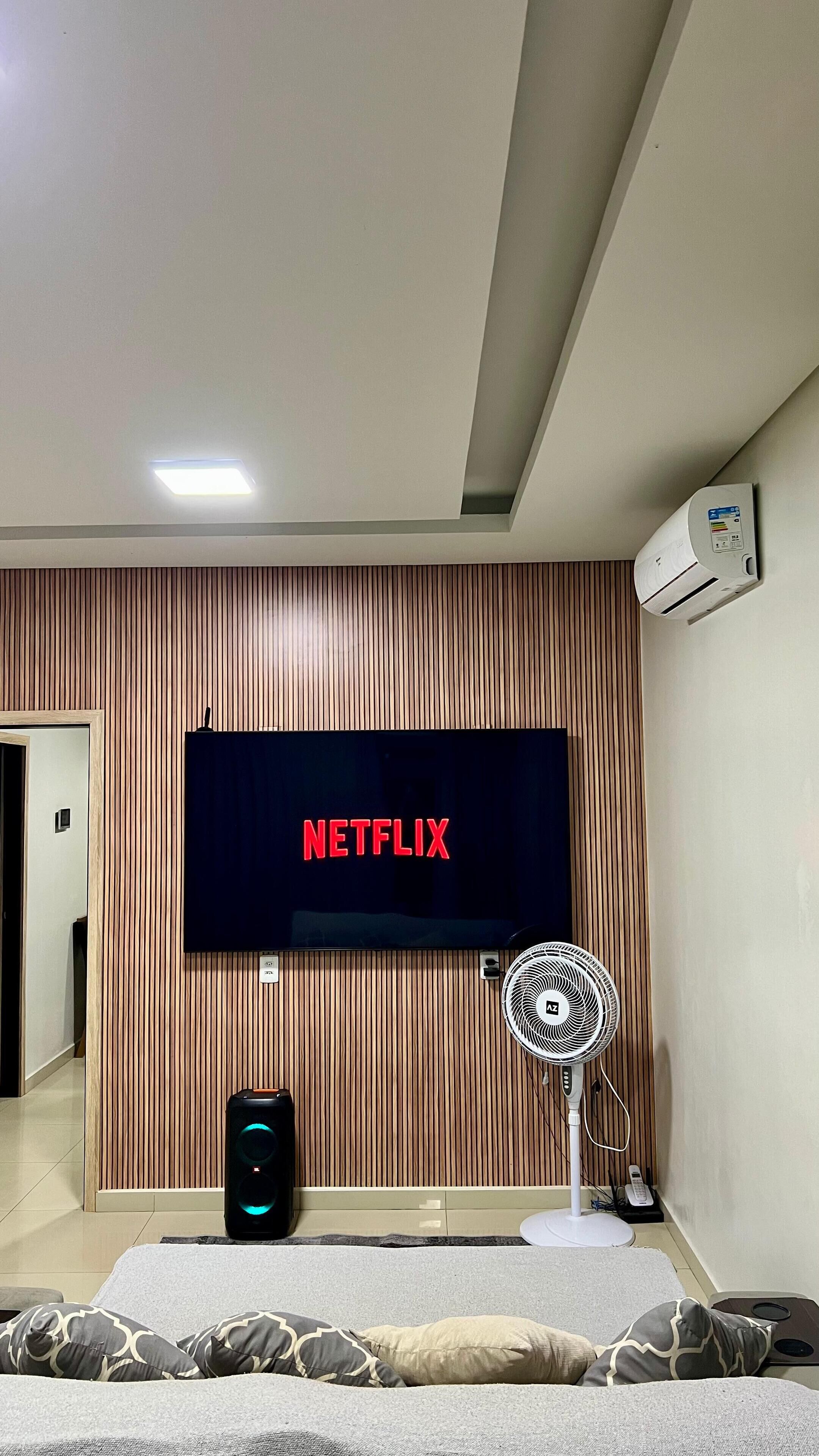 Een moderne woonkamer met een tv aan de muur waarop het Netflix logo te zien is, een staande ventilator en een speaker.