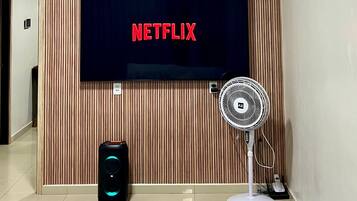 Een moderne woonkamer met een tv aan de muur waarop het Netflix logo te zien is, een staande ventilator en een speaker.