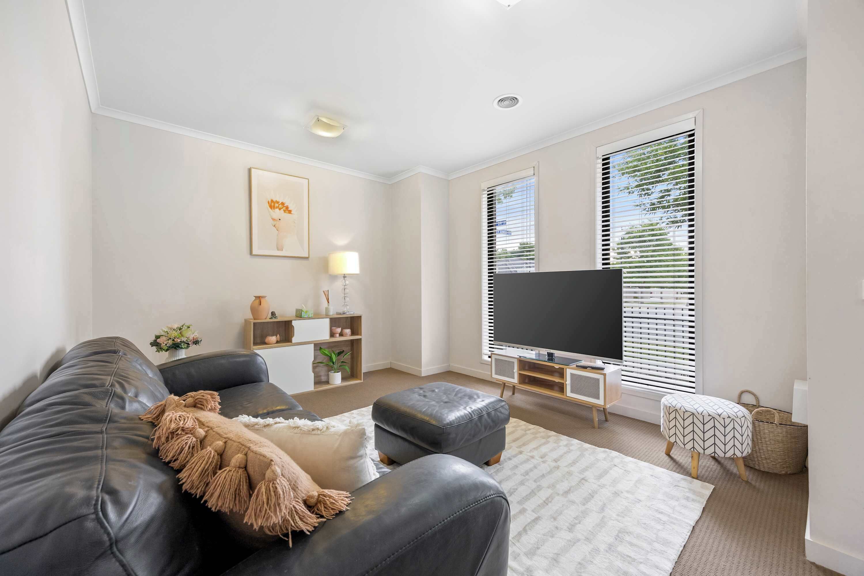 A modern living room with a leather sofa, ottoman, and a flat-screen TV. There is a small side table with a lamp, a bookshelf, and a window with blinds.