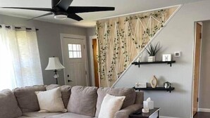 A modern living room with a sectional sofa, a round ottoman, a ceiling fan, and a staircase with a decorative wall. - LVR's 3 Sister's Cape (Lewiston)
