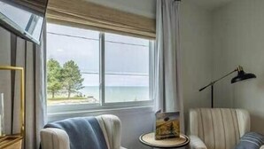 Una habitación con un gran ventanal que da a una masa de agua, dos sillones, una mesita con un libro y una lámpara de pie.