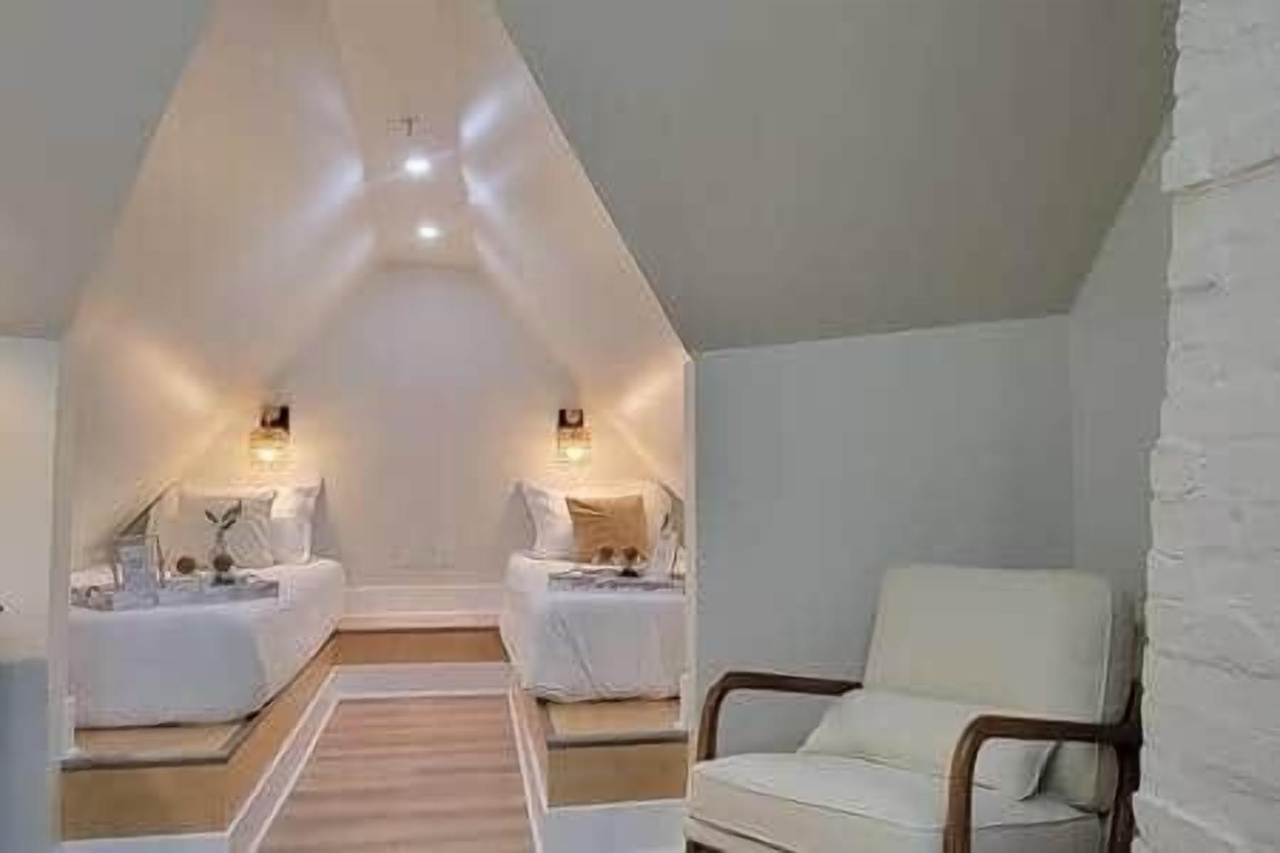 Een gezellige, goed verlichte kamer met een schuine kap, twee nachtkastjes met lampen, een comfortabele stoel en een houten vloer.