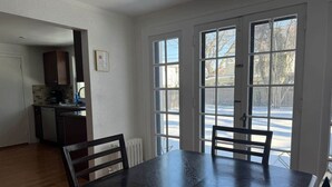 A dining area with a dark wooden table and chairs, a kitchen in the background, and large windows with a view of a snowy landscape.