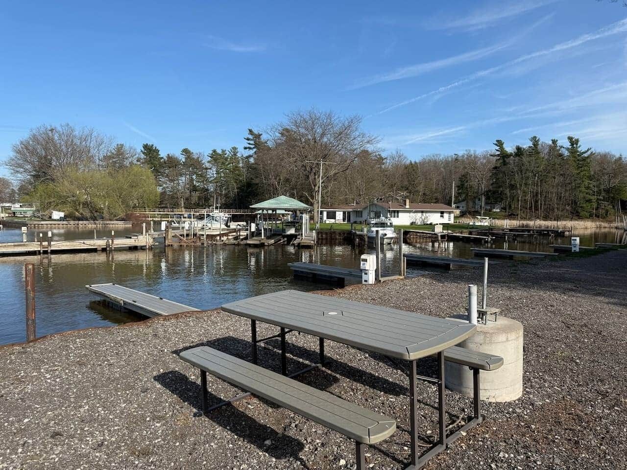 A lakeside picnic area with a table and benches, a dock, and boats in the background.