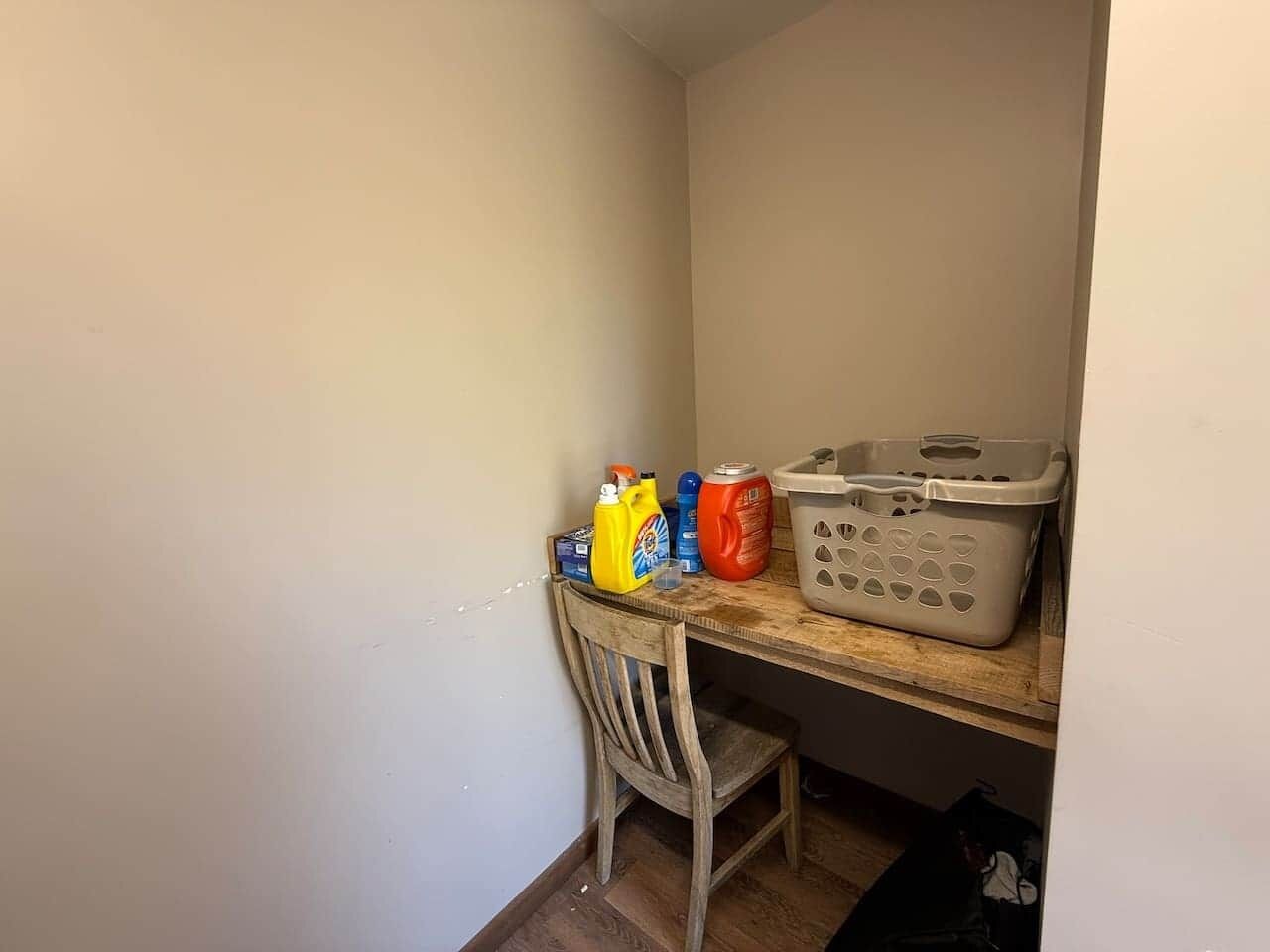 A small kitchen area with a wooden chair, a table with cleaning supplies, and a laundry basket.