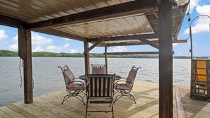 A covered outdoor seating area with a table and four chairs, overlooking a calm lake. - Hot Tub, Fire Pit, Kayak,Water Slide, Pet Friendly (Bracey)