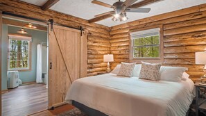 A log cabin bedroom with a bed, two bedside lamps, a ceiling fan, and a window. - Hot Tub, Fire Pit, Kayak,Water Slide, Pet Friendly (Bracey)