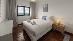 A neatly arranged bedroom with a large bed, a bedside table with a lamp, a window with curtains, and a view of the sea. - Pérola do Mar - Atlantic Escape (Caldas da Rainha)