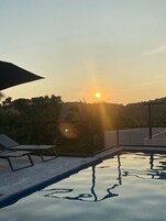 Una piscina con el sol poniéndose en el fondo, rodeada de vegetación y un cielo despejado.