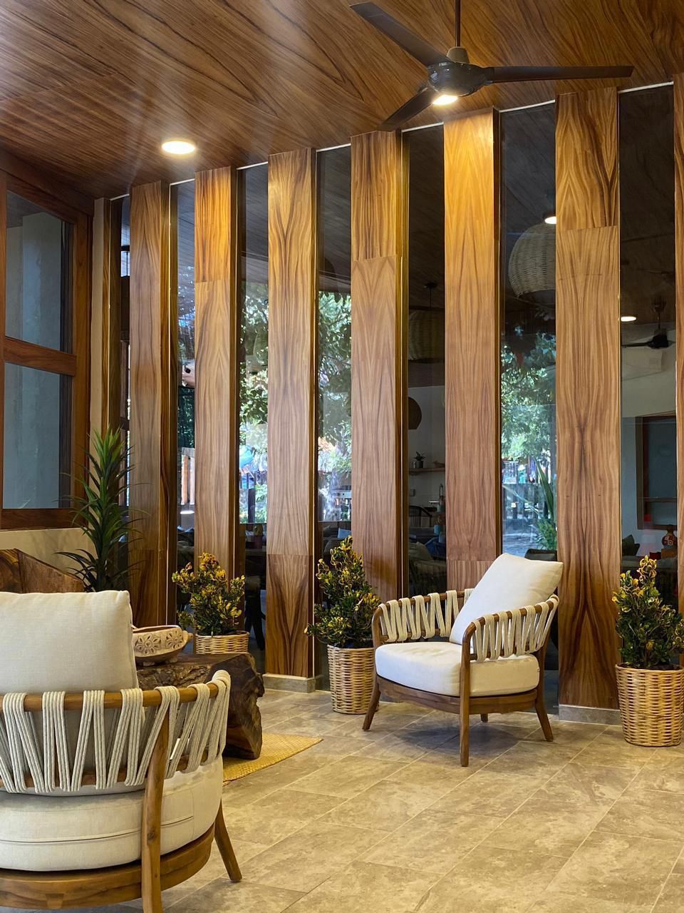 A room with a wooden ceiling, a ceiling fan, and a large window with vertical wooden panels. There are two chairs and a small table.