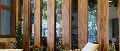 Une pièce avec un plafond en bois, un ventilateur de plafond et une grande fenêtre dotée de panneaux de bois verticaux. On y trouve deux chaises et une petite table.
