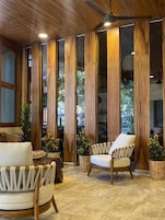 A room with a wooden ceiling, a ceiling fan, and a large window with vertical wooden panels. There are two chairs and a small table.